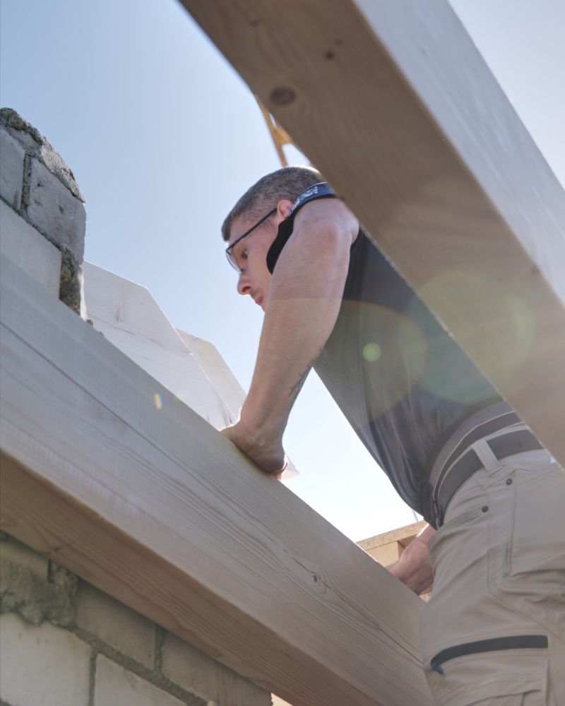 Handwerker auf dem Dach legt einen neuen Holzbalken auf das Mauerwerk, von unten gegen den Himmel fotografiert.