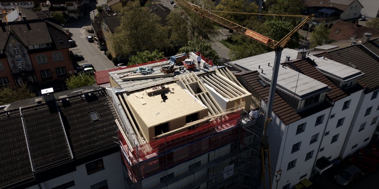 Luftaufnahme einer laufenden Aufstockung: Auf einem eingerüsteten Mehrfamilienhaus entsteht ein neues Dachgeschoss in Holzbauweise, daneben ein Baukran.