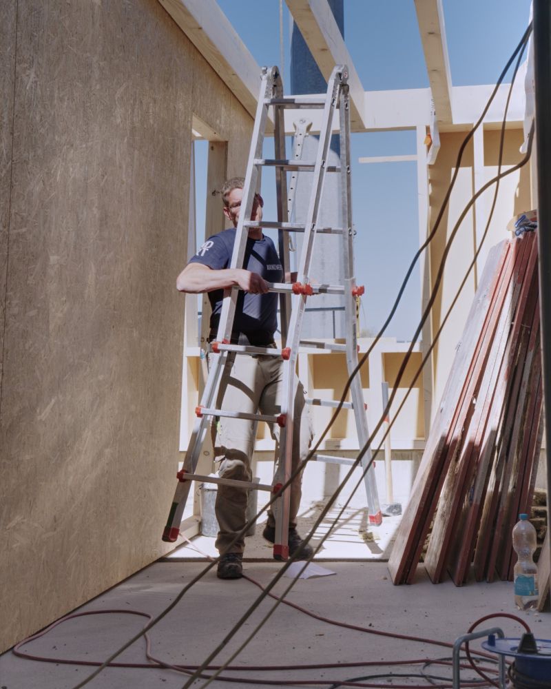 Handwerker im Handwertig-T-Shirt trägt eine Leiter durch das neue Dachgeschoss, darüber die offenen Deckenbalken.