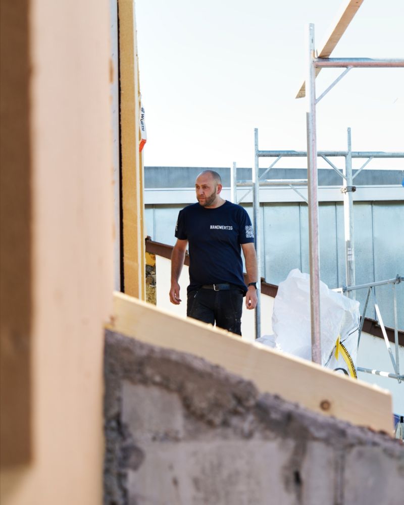 Mitarbeiter im Handwertig-T-Shirt auf der Baustelle, im Vordergrund Mauerwerk und ein neuer Holzbalken.