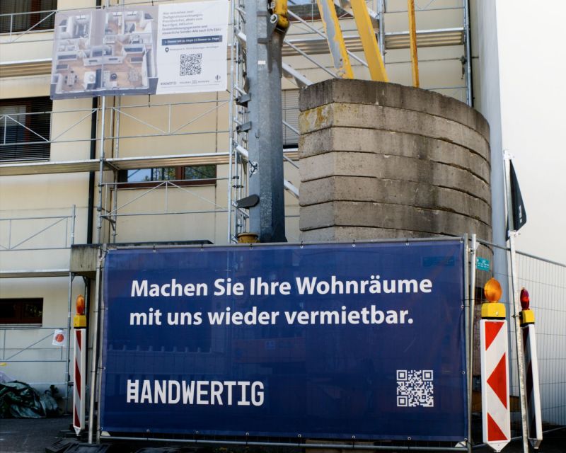 Eingerüstetes Mehrfamilienhaus mit Bauschild „Hier entstehen zwei Dachgeschosswohnungen“, davor ein Bauzaun mit dem Banner „Machen Sie Ihre Wohnräume mit uns wieder vermietbar.“
